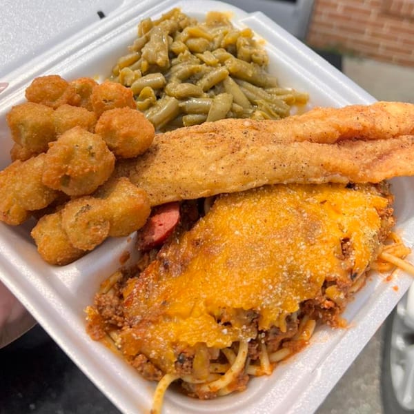 A soul food meal with fried fish, fried okra, green beans, and baked spaghetti in a to-go container.